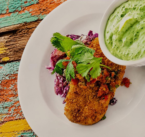 Is there anything better than a crispy, breaded fried chicken breast? I’ll answer that for you: No. There’s no secret to achieving it, but you do need to master proper technique and add a little creativity. Do it right, you’ll end up with a cutlet that’s light and crunchy on the outside and tender and juicy on the inside. Get it wrong, and you’ll be dining on bland, tasteless chicken with a soggy, uninspired coating that slides right off the meat. It will be edible, but most likely boring and average. And who wants ho-hum when you can achieve flavorful, crispy perfection? So, for this month’s VDP recipe, I’m giving you the skinny on how to solve this age-old fried chicken dilemma. For starters, you’ll want to slice the chicken breast neatly in half horizontally with a sharp knife. If the breast is too thick, it will never cook all the way through before the outside burns. If it’s too thin, you run the risk of overcooking and drying out the meat before you’ve achieved the perfect crust. After you’ve sliced the breast, season the paillards with salt and pepper and let them rest for about 10 minutes so the flavor can work its way into the meat. Next up is the breading. The traditional three-step technique is the standard fried chicken go-to. But here we’re going to streamline the process by combining the egg and the flour to make a slurry that will act as a glue, giving the crumbs something to adhere to. Without it, the crumbs will slide right off the meat. Once the chicken has been dredged in the egg-flour mixture, coat the breasts in the crumbs and press lightly to help them adhere. Now, here’s your chance to really bring home the flavor and let your creativity shine. The most basic option for the crumb coating is traditional breadcrumbs, but you can also use panko, breakfast cereal, crushed pretzels, nuts, seeds, crackers, instant ramen, or my favorite, snack chips. Potato chips are great, but I’m especially partial to corn chips. They give this dish that sweet, earthy south-of-the-border flair. After the chicken breasts are covered in the crumb coating, let them rest for another 15 minutes. This gives the coating time to really adhere to the meat and helps keep it in place when you fry the chicken. A quick fry in a cast iron skillet or Dutch oven gives you a golden, crispy crust and consistent, delicious results every time. Served with coleslaw and black beans and you’ve got a definite dinnertime win! If you have not joined the Virtual Dinner Party but would like to dine with us, please prepare this recipe—or any other—and share it with your family, friends, and neighbors. It is our group’s firm belief that by preparing something good to eat and sharing it with those we love, we are participating in one of life’s greatest joys. The connection, community, and goodwill that come from it fill us with joy, hope, and gratitude. I hope you all enjoy it as much as I do. Thanks for coming to the party, and cook on! – Vic